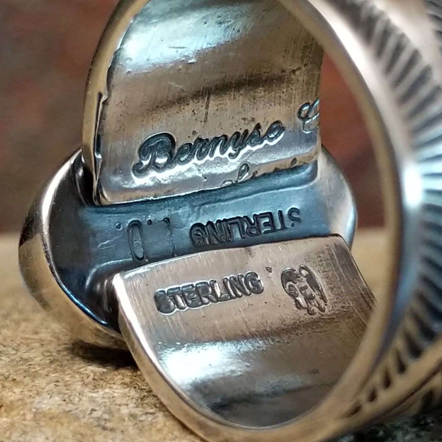 A large, oval Golden Hills turquoise ring by Bernyse Chavez & Lowell Draper, featuring a sky-blue stone with brown matrix veining, set in a wide sterling silver band with a symmetrical sun-ray pattern. The ring rests on a textured sandstone surface with a warm, earthy red background.