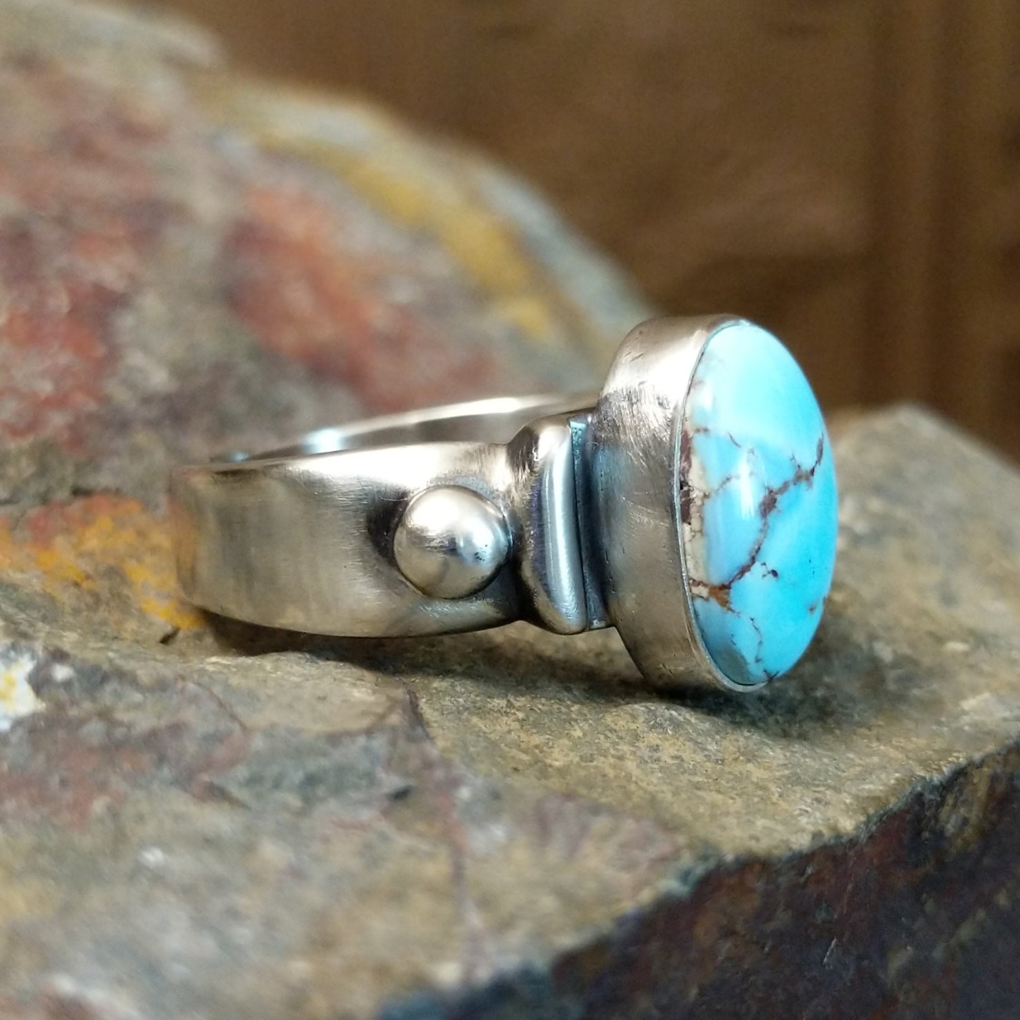 Silver ring with a smooth bezel holding an oval turquoise stone and raised silver detailing on the band.