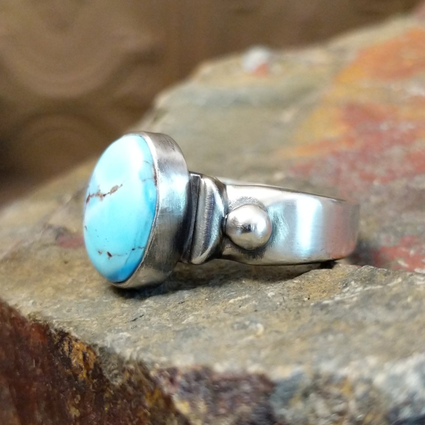 Silver ring with a smooth bezel holding an oval turquoise stone and raised silver detailing on the band.