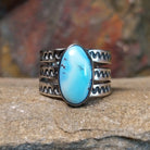 An oval Golden Hills turquoise ring by Raymond Coriz, featuring a sky-blue stone with brown matrix veining, set in a sterling silver band with three layered sections, each with a zigzag pattern. The ring rests on a textured sandstone surface with a warm, earthy red background.