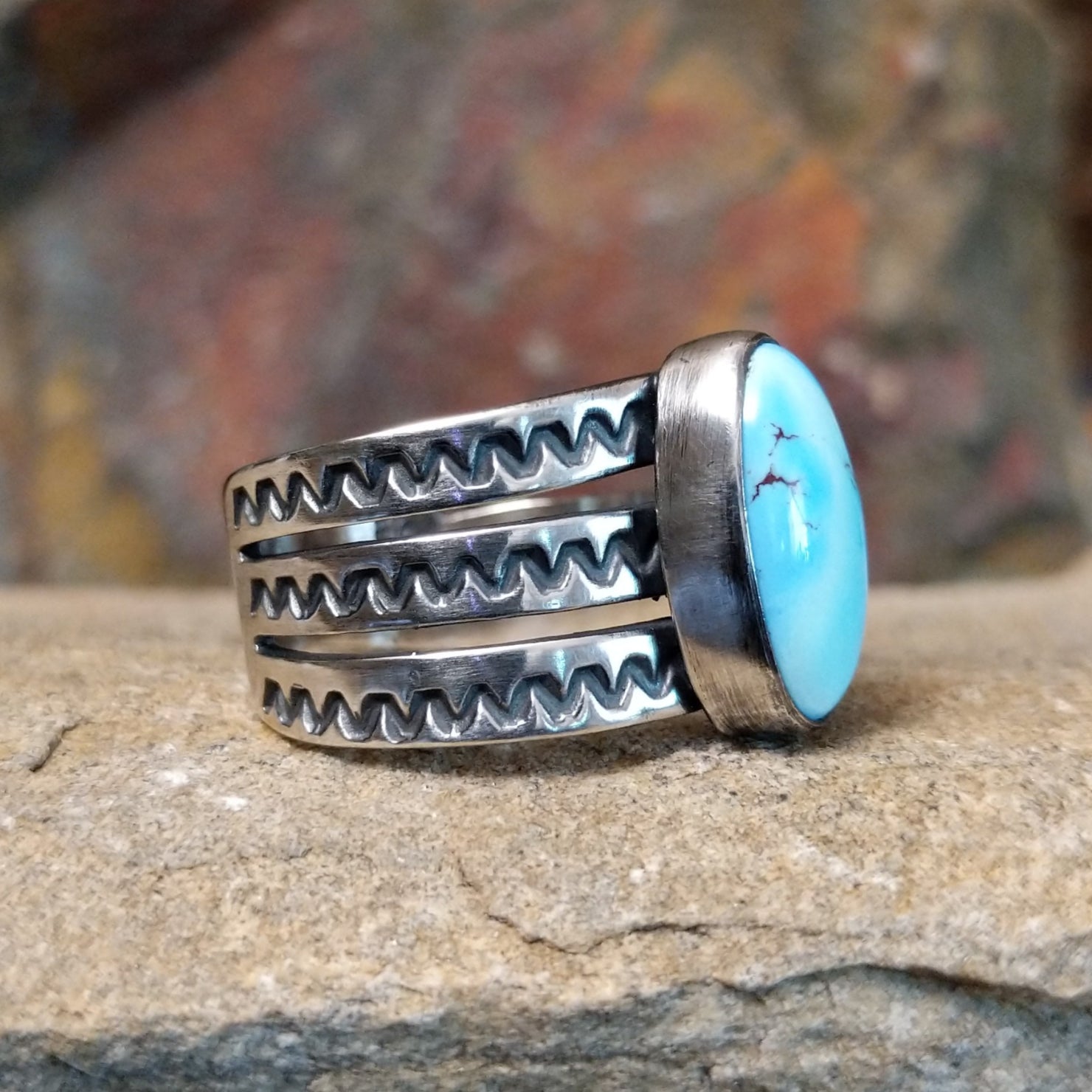 An oval Golden Hills turquoise ring by Raymond Coriz, featuring a sky-blue stone with brown matrix veining, set in a sterling silver band with three layered sections, each with a zigzag pattern. The ring rests on a textured sandstone surface with a warm, earthy red background.
