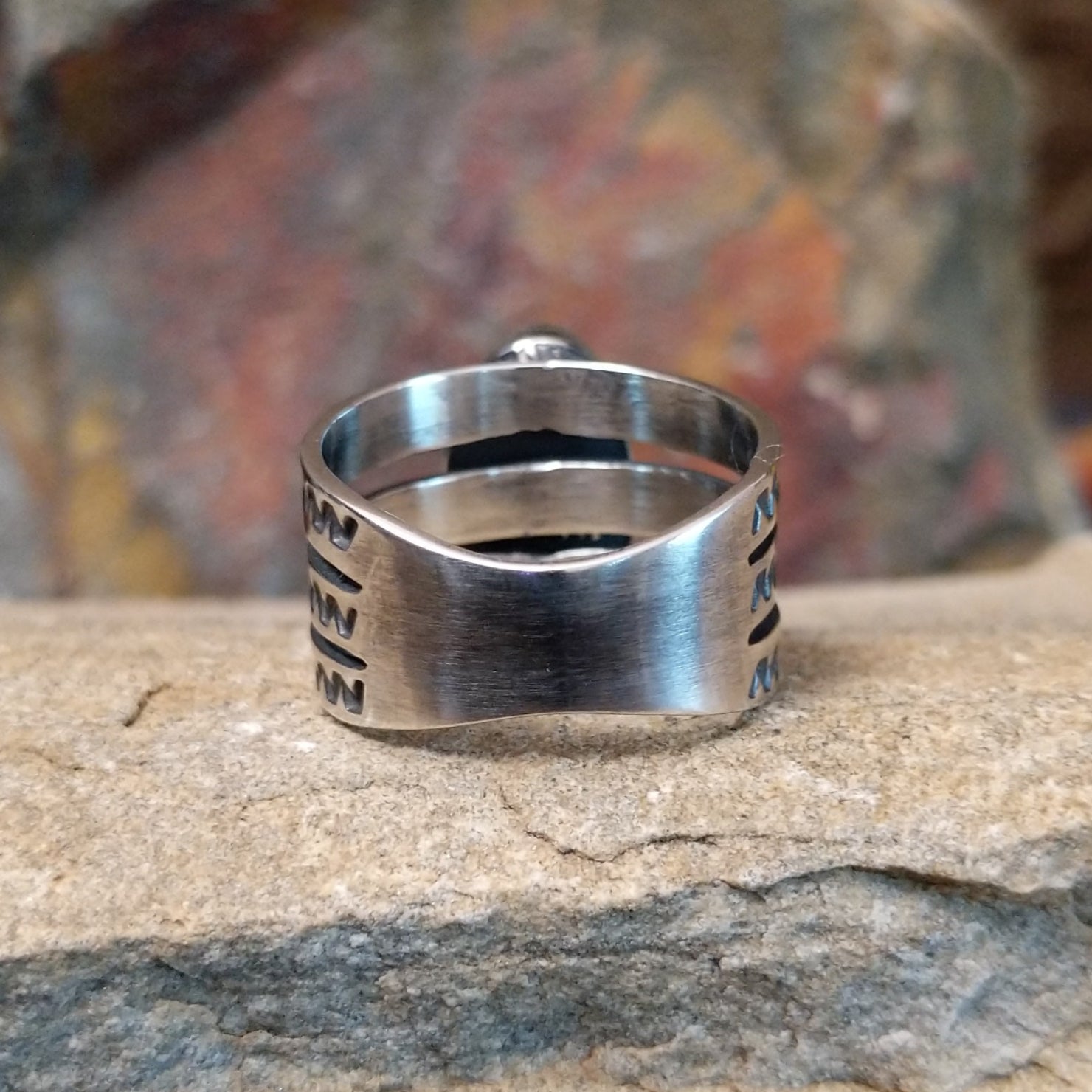 An oval Golden Hills turquoise ring by Raymond Coriz, featuring a sky-blue stone with brown matrix veining, set in a sterling silver band with three layered sections, each with a zigzag pattern. The ring rests on a textured sandstone surface with a warm, earthy red background.
