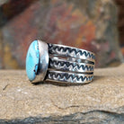 An oval Golden Hills turquoise ring by Raymond Coriz, featuring a sky-blue stone with brown matrix veining, set in a sterling silver band with three layered sections, each with a zigzag pattern. The ring rests on a textured sandstone surface with a warm, earthy red background.
