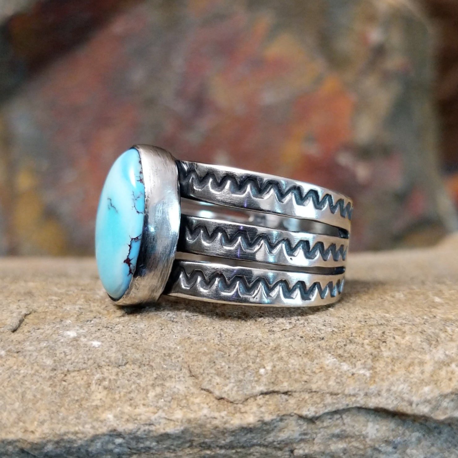 An oval Golden Hills turquoise ring by Raymond Coriz, featuring a sky-blue stone with brown matrix veining, set in a sterling silver band with three layered sections, each with a zigzag pattern. The ring rests on a textured sandstone surface with a warm, earthy red background.
