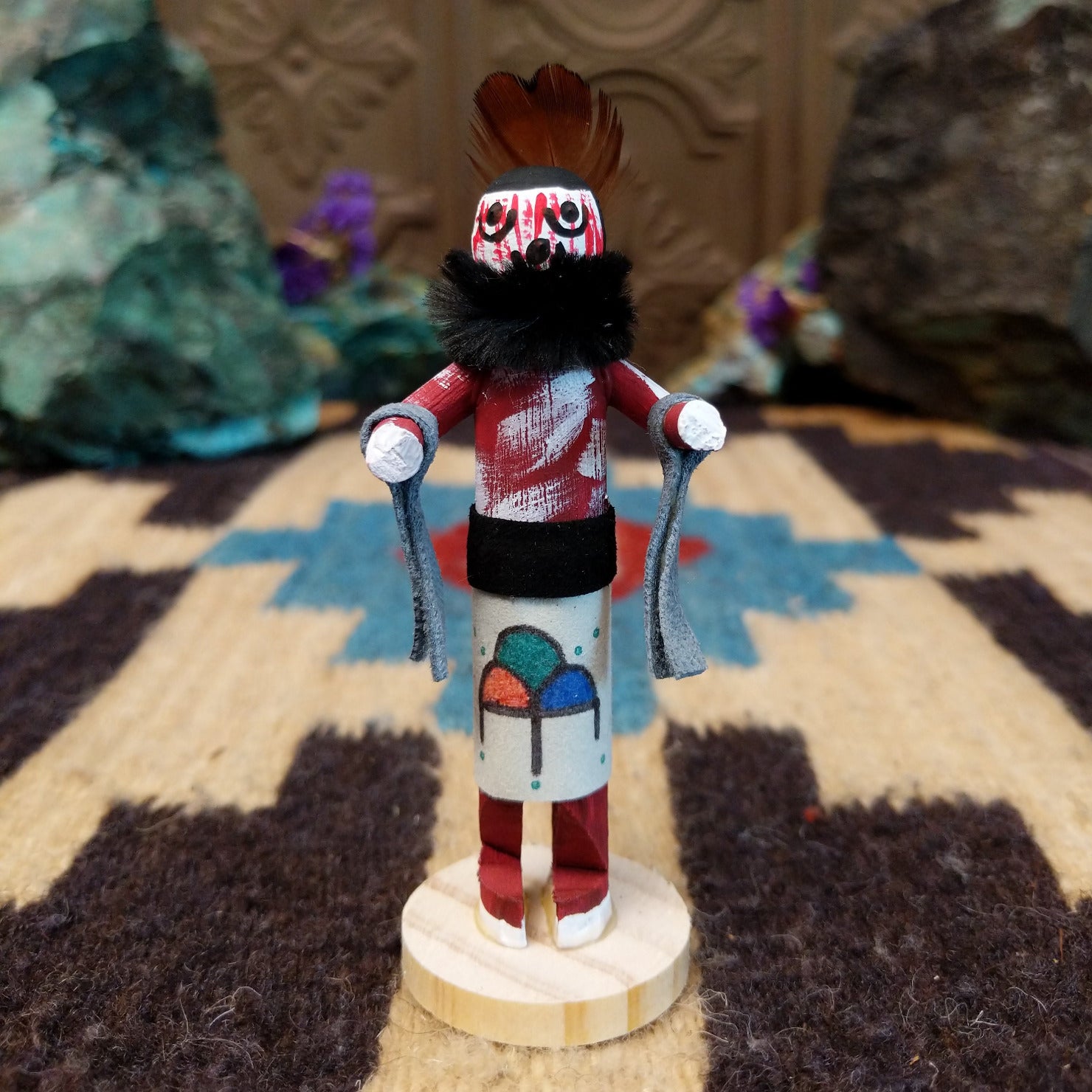 Mini grandmother kachina by Rosalind Grey with red body, black fur collar, feathered headpiece, and white geometric-patterned garment.