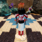 Mini grandmother kachina by Rosalind Grey with red body, black fur collar, feathered headpiece, and white geometric-patterned garment.