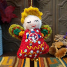 This whimsical Green-Winged Angel Wool Ornament features golden yellow hair, a smiling white face, embroidered green wings, a red dress with colorful stitching and silver sequins, dangling gold bells, and a gold hanging cord.