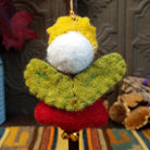 This whimsical Green-Winged Angel Wool Ornament features golden yellow hair, a smiling white face, embroidered green wings, a red dress with colorful stitching and silver sequins, dangling gold bells, and a gold hanging cord; back side view.