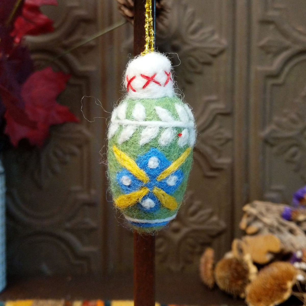 This delightful Green Bulb Wool Ornament by Ganz features a soft green wool body decorated with blue and yellow floral accents, white leaf patterns, a fluffy white top with red stitching, and a gold hanging cord.