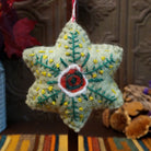 This distinctive Green Star Wool Ornament by Ganz features a soft sage green six-pointed wool star with a red floral centerpiece, embroidered green leafy branches, yellow accents, and a red and white twisted hanging cord.
