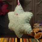 This distinctive Green Star Wool Ornament by Ganz features a soft sage green six-pointed wool star with a red floral centerpiece, embroidered green leafy branches, yellow accents, and a red and white twisted hanging cord; back side view.
