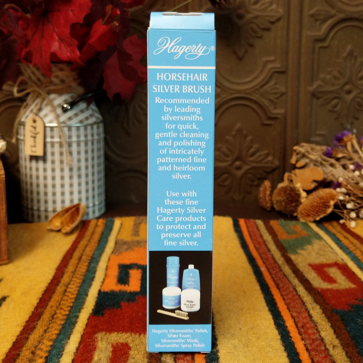 A tall blue and black box of Hagerly Horsehair Silver Brush stands upright on a patterned rug in a styled indoor setting with decorative dried flowers and a textured brown wall behind it.