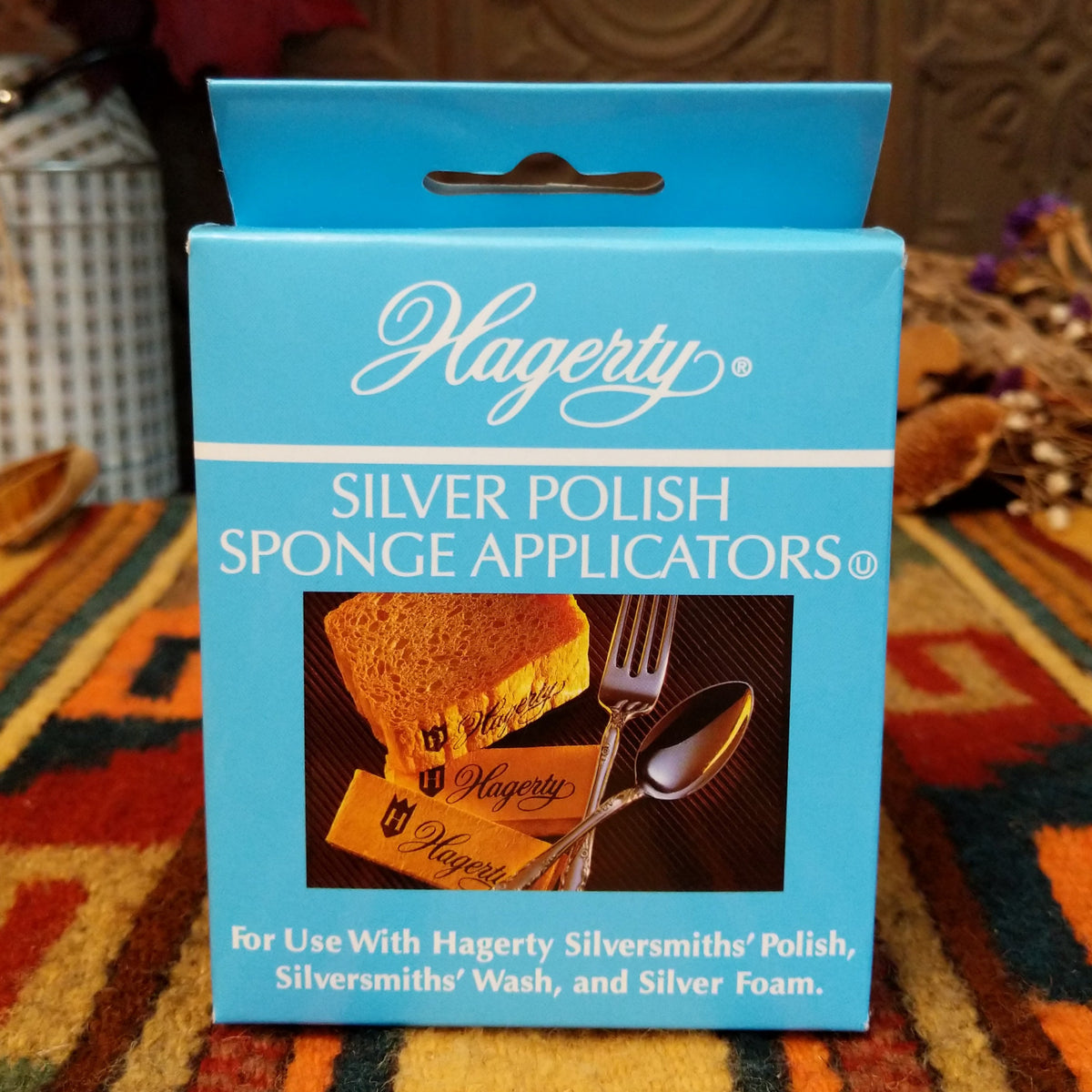 A blue box of Hagerty Silver Polish Sponge Applicators, shown standing upright on a patterned rug with household items blurred in the background.