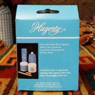 A light blue Hagerty product box standing on a patterned orange and brown rug, with a dark wood carved backdrop and dried floral decor in the background.