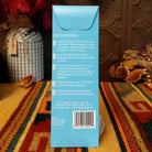 The back of a light blue product box standing upright on a patterned rug, with printed instructions and a barcode visible. Decorative dried flowers and a dark wood wall are in the background.