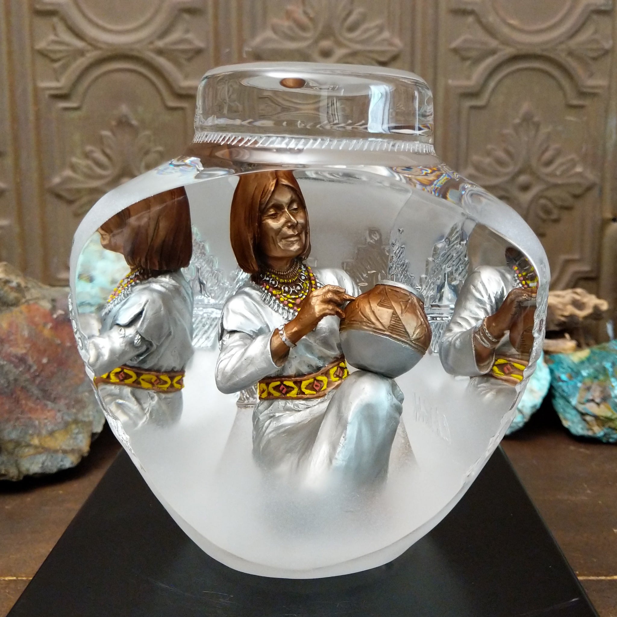 An Indigenous woman decorating a traditional pottery vessel, enclosed in a clear Lucite sculpture that creates mirrored reflections. The figure wears a silver-toned dress with colorful beaded jewelry.