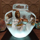 An Indigenous woman decorating a traditional pottery vessel, enclosed in a clear Lucite sculpture that creates mirrored reflections. The figure wears a silver-toned dress with colorful beaded jewelry; lower light display.
