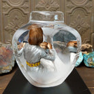 An Indigenous woman decorating a traditional pottery vessel, enclosed in a clear Lucite sculpture that creates mirrored reflections. The figure wears a silver-toned dress with colorful beaded jewelry; rear view.