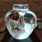 An Indigenous woman decorating a traditional pottery vessel, enclosed in a clear Lucite sculpture that creates mirrored reflections. The figure wears a silver-toned dress with colorful beaded jewelry; lower light rear display.