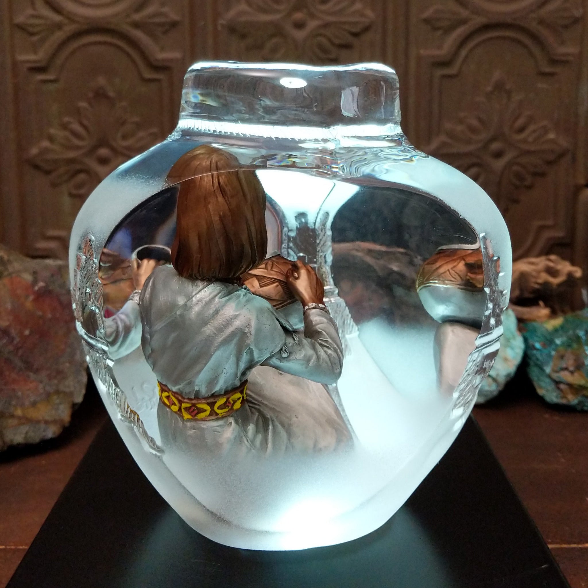 An Indigenous woman decorating a traditional pottery vessel, enclosed in a clear Lucite sculpture that creates mirrored reflections. The figure wears a silver-toned dress with colorful beaded jewelry; lower light rear display.