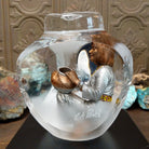 An Indigenous woman decorating a traditional pottery vessel, enclosed in a clear Lucite sculpture that creates mirrored reflections. The figure wears a silver-toned dress with colorful beaded jewelry; side view with artisan signature and production number.