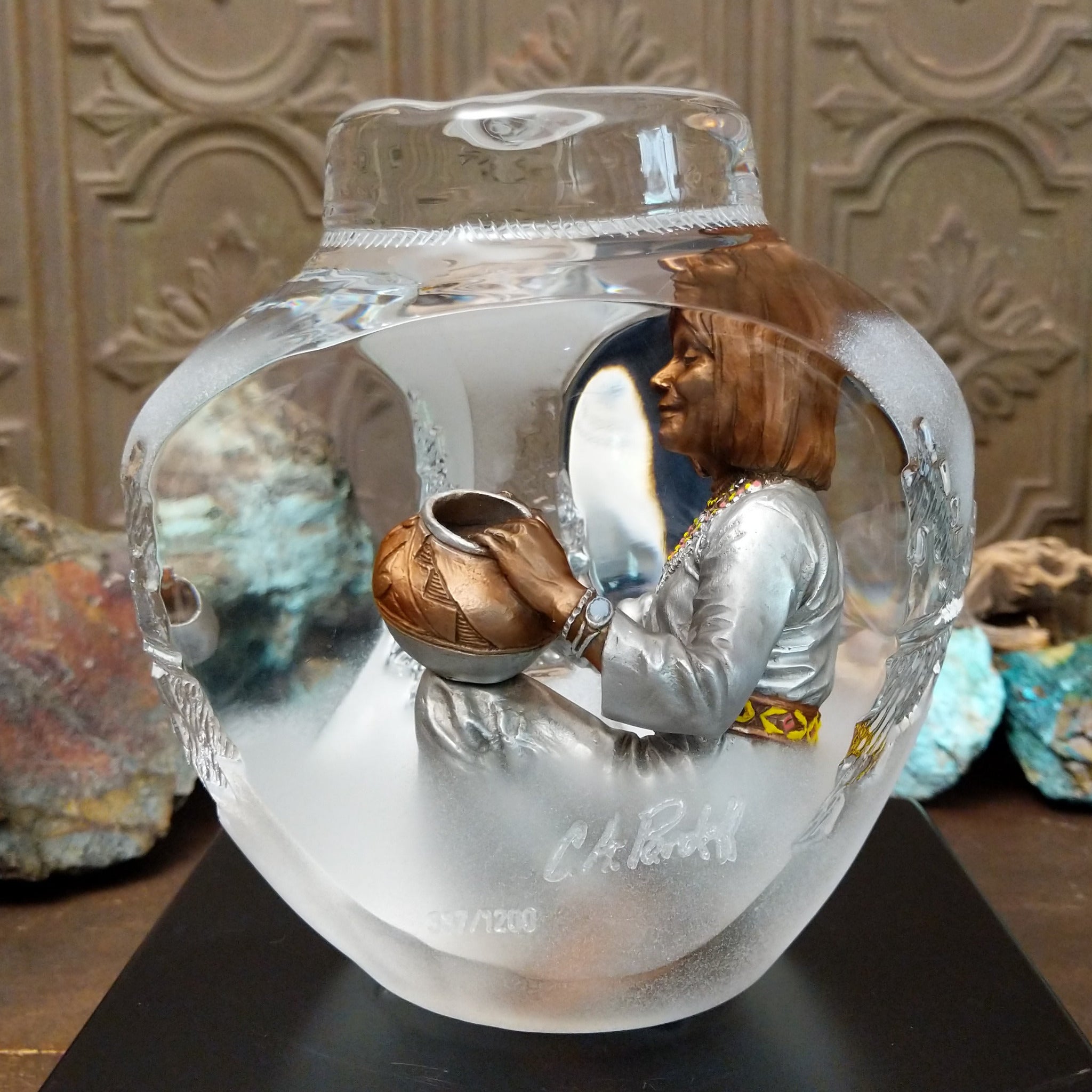 An Indigenous woman decorating a traditional pottery vessel, enclosed in a clear Lucite sculpture that creates mirrored reflections. The figure wears a silver-toned dress with colorful beaded jewelry; side view with artisan signature and production number.