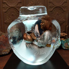 An Indigenous woman decorating a traditional pottery vessel, enclosed in a clear Lucite sculpture that creates mirrored reflections. The figure wears a silver-toned dress with colorful beaded jewelry; lower light view with artisan signature and production number.
