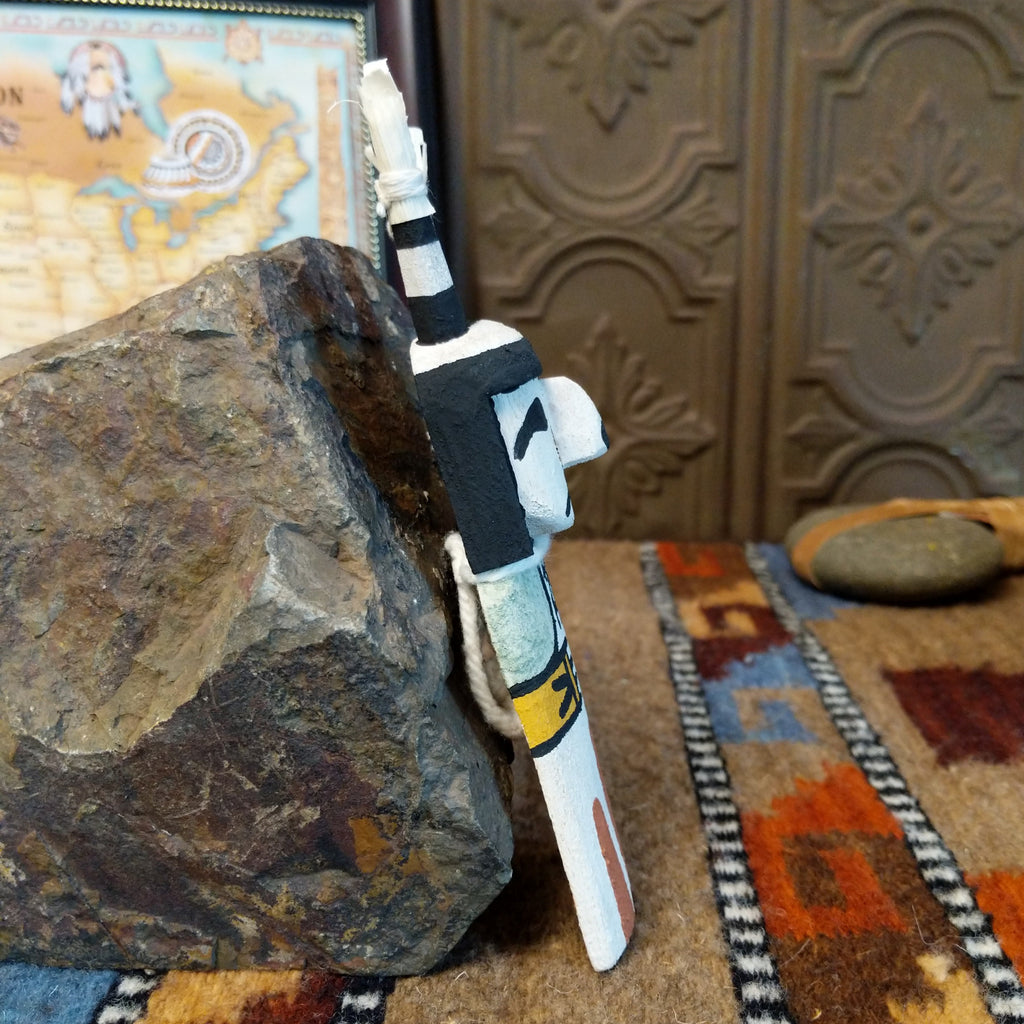 Wood carved into a clown kachina painted with black and white paint with pops of color and features corn husk horns.