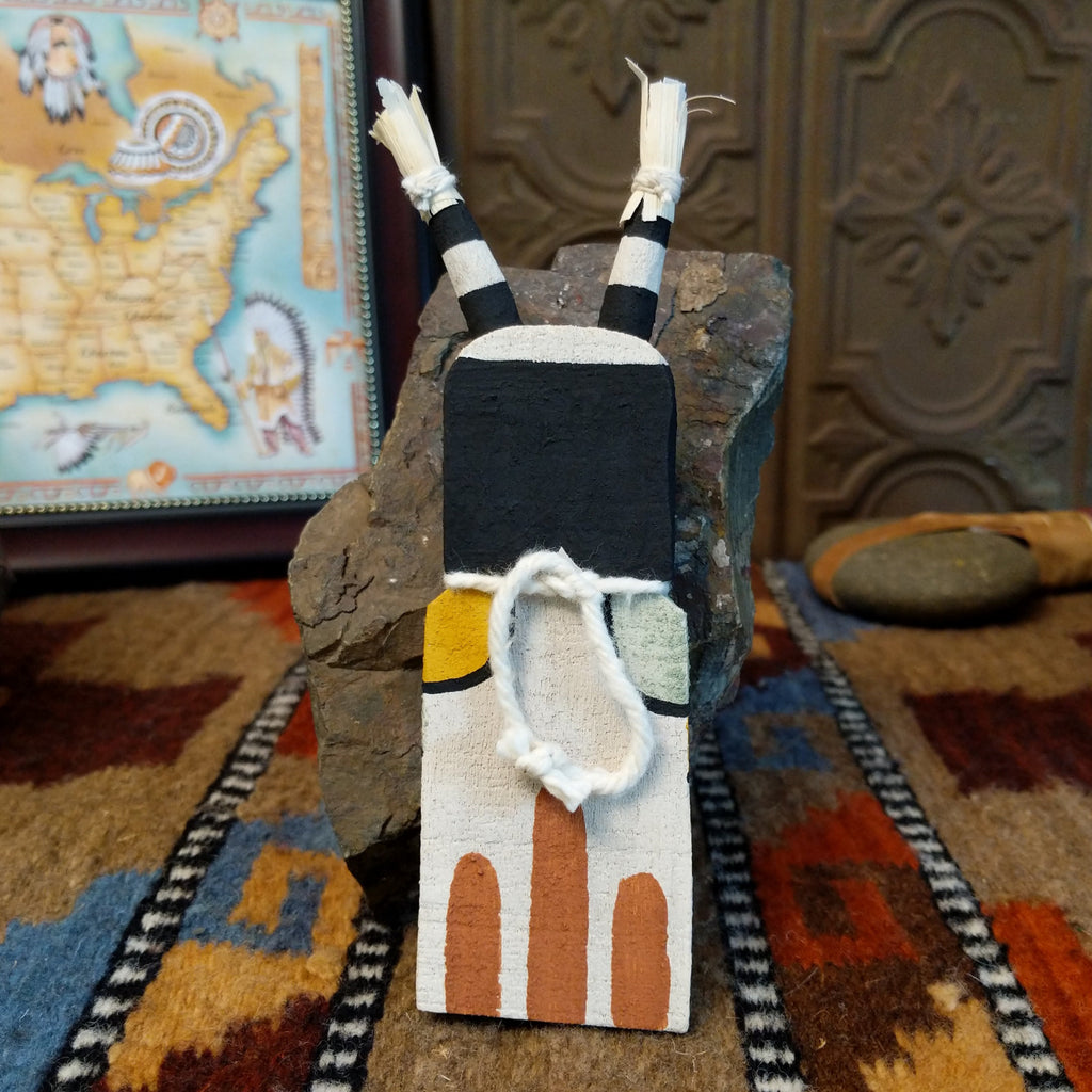 Wood carved into a clown kachina painted with black and white paint with pops of color and features corn husk horns.