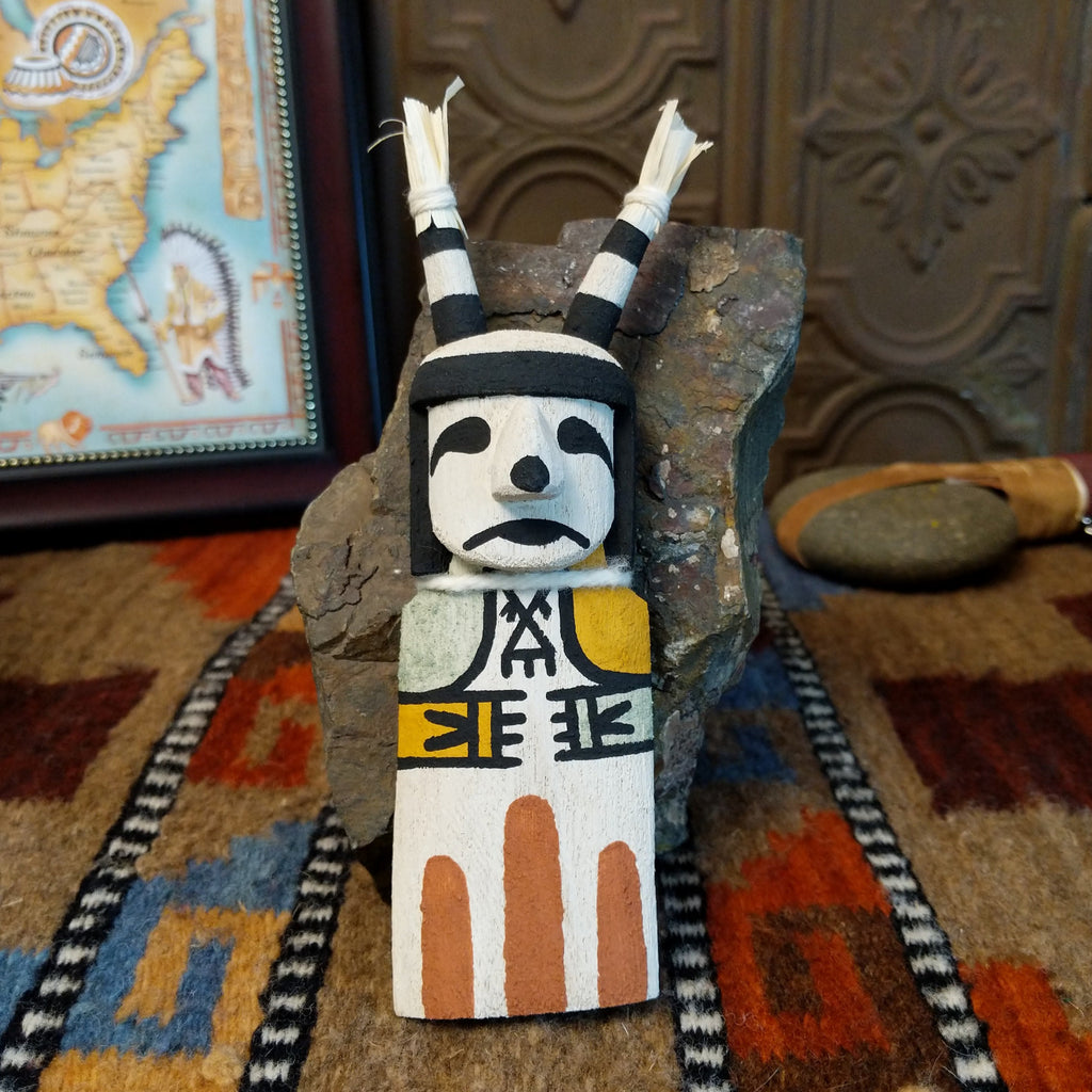 Wood carved into a clown kachina painted with black and white paint with pops of color and features corn husk horns.