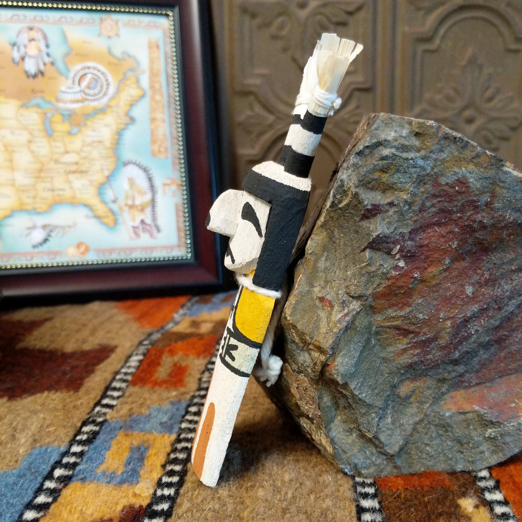 Wood carved into a clown kachina painted with black and white paint with pops of color and features corn husk horns.