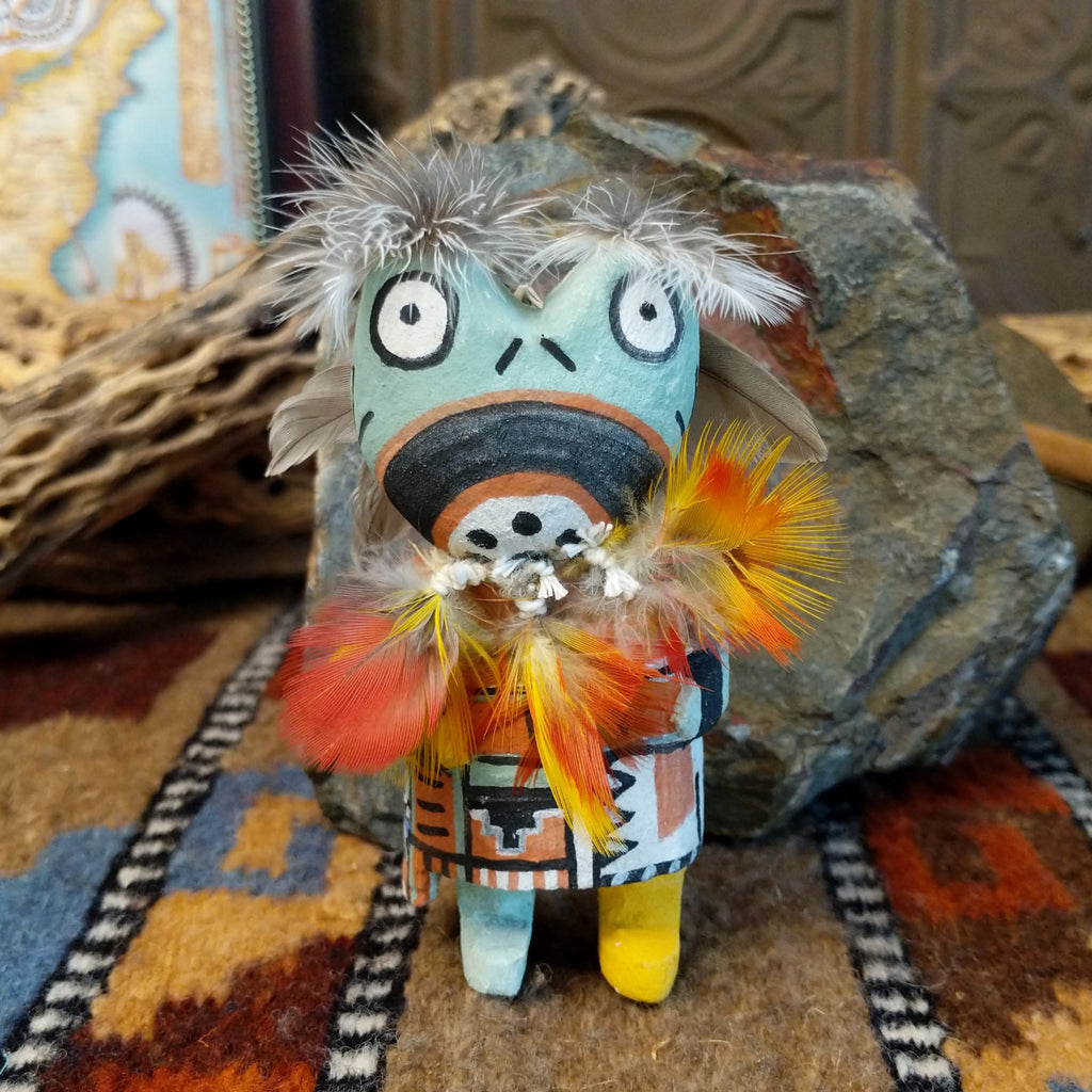 Wood carved into a frog kachina doll, painted with vibrant colors and feathers adorning its' body.