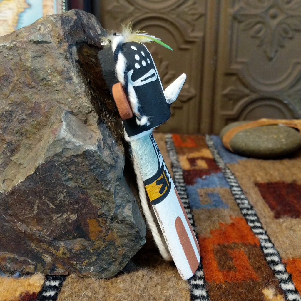 Wood carved into a kokopelli kachina with black, white, orange, and yellow paint featuring feathers at the top of the head.