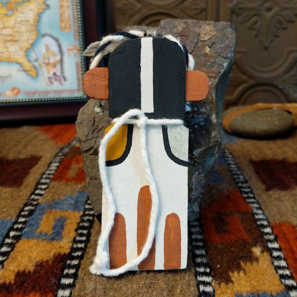 Wood carved into a kokopelli kachina with black, white, orange, and yellow paint featuring feathers at the top of the head and string tied at the back.