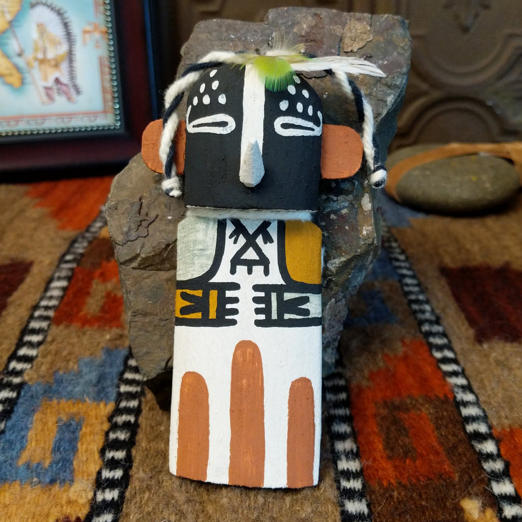 Wood carved into a kokopelli kachina with black, white, orange, and yellow paint featuring feathers at the top of the head.