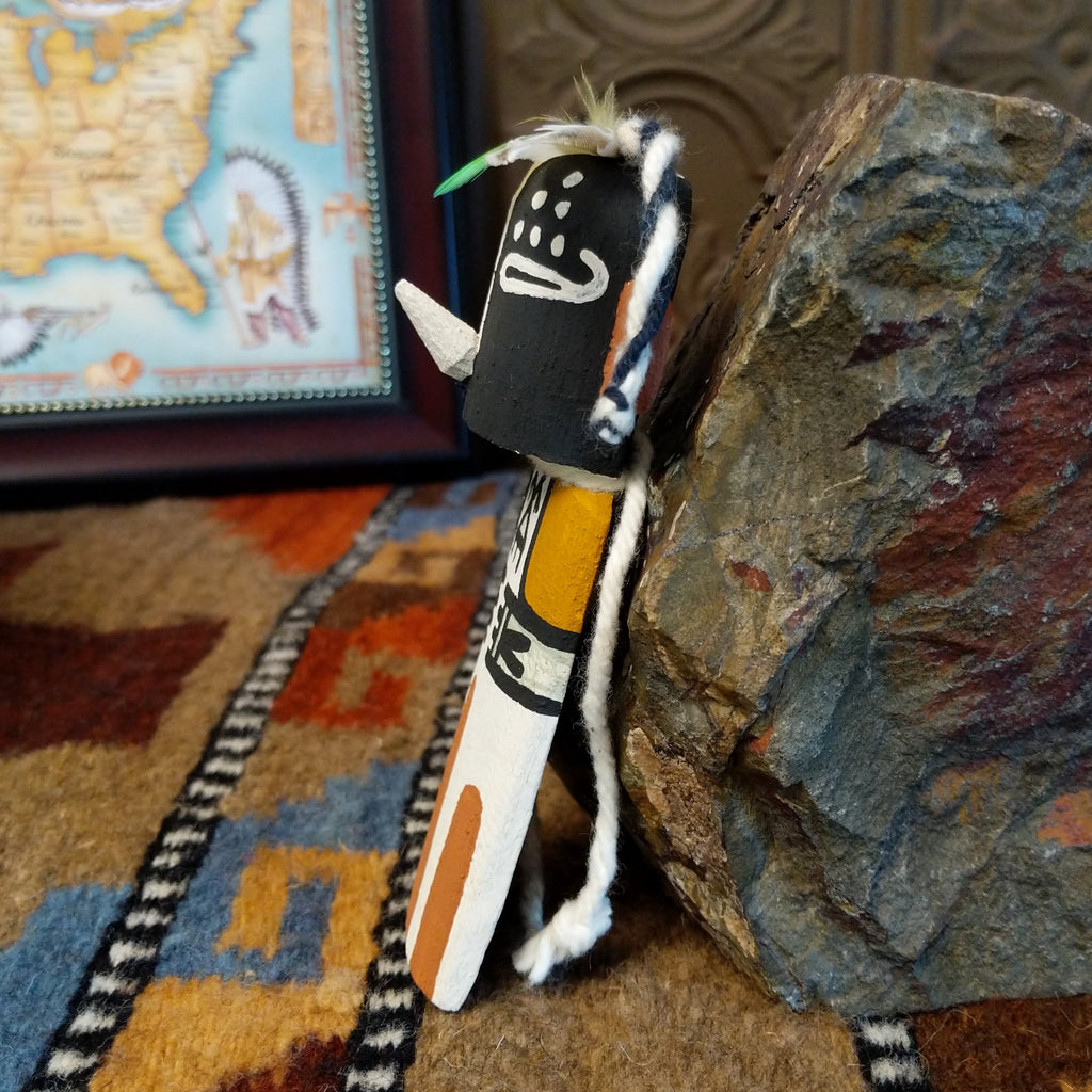 Wood carved into a kokopelli kachina with black, white, orange, and yellow paint featuring feathers at the top of the head.