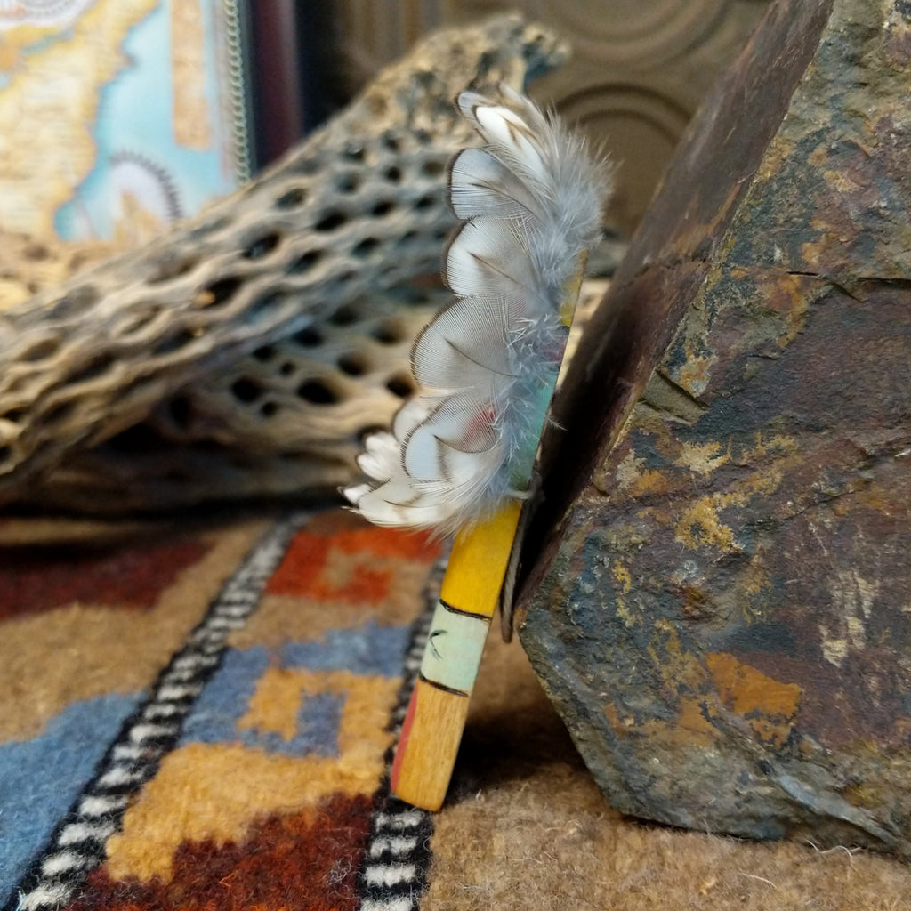 Wood carved into a sunface, painted with vibrant colors and features feathers.