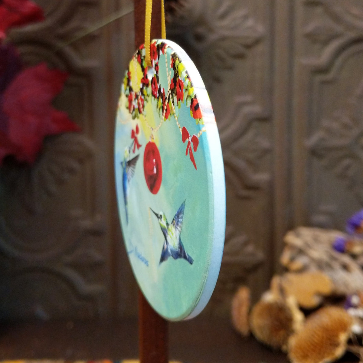 A round hanging ornament with a light blue background, painted hummingbirds, red butterflies, floral accents, and a red hanging loop, shown hanging against a dark carved wooden backdrop.