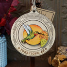 Hummingbird Sunset Wood Ornament featuring a colorful green hummingbird, orange desert flowers, sunset background, engraved circular wooden frame, “Tombstone, AZ” lettering, and natural twine hanging loop.