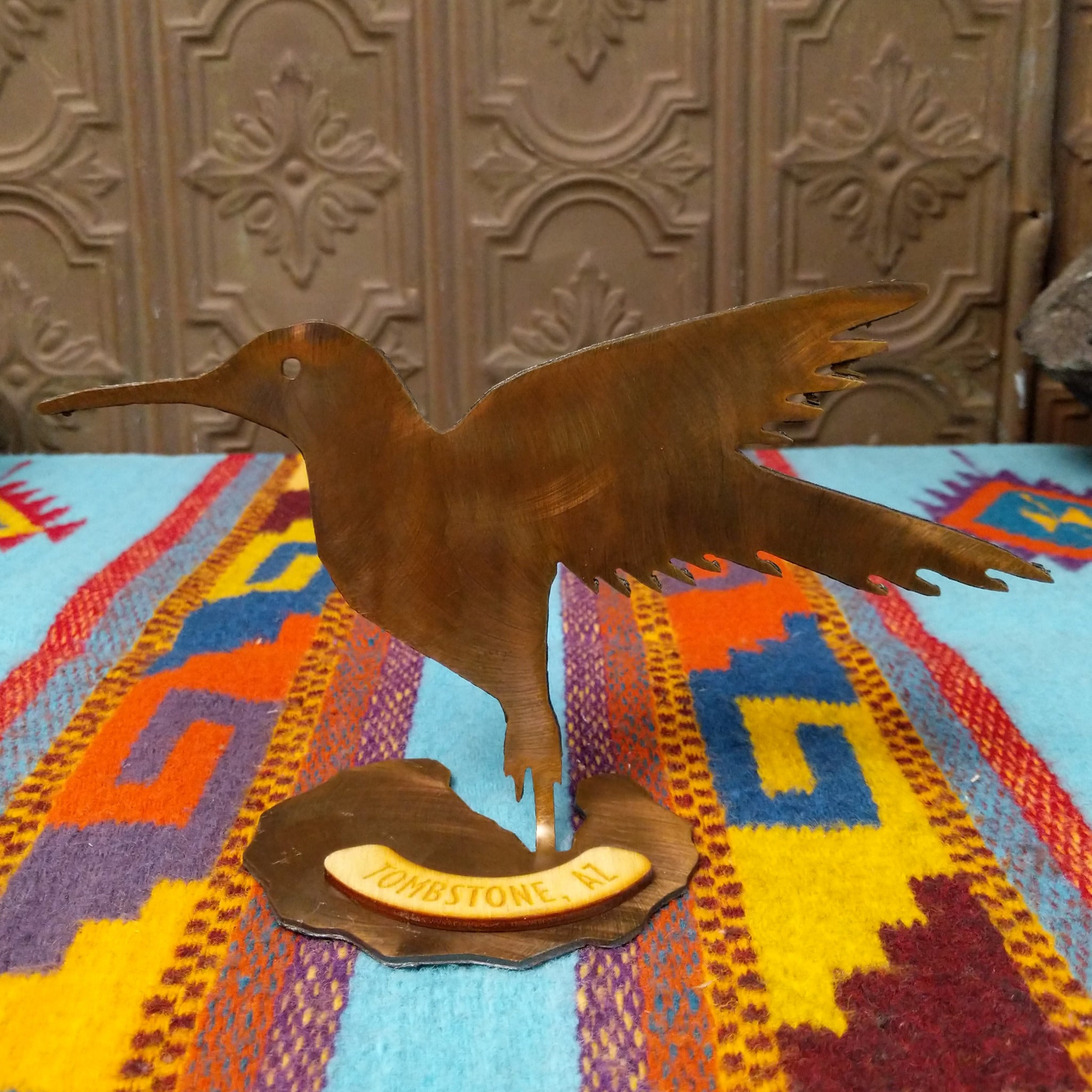 Handcrafted Crooks Design metal hummingbird tabletop sculpture with outstretched wings, detailed feather cutouts, a warm burnished finish, and a decorative base marked Tombstone, AZ.