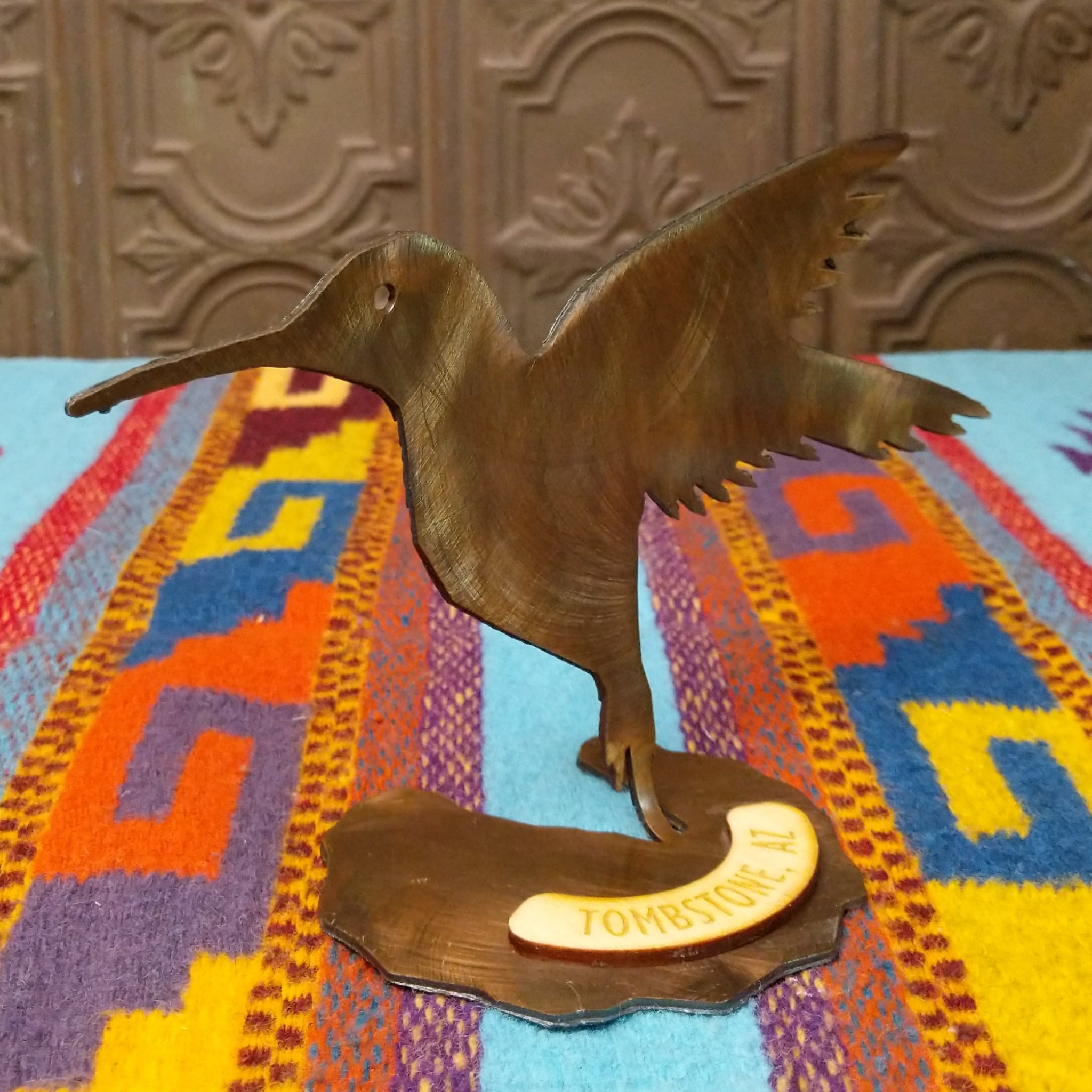Handcrafted Crooks Design metal hummingbird tabletop sculpture with outstretched wings, detailed feather cutouts, a warm burnished finish, and a decorative base marked Tombstone, AZ; side view.