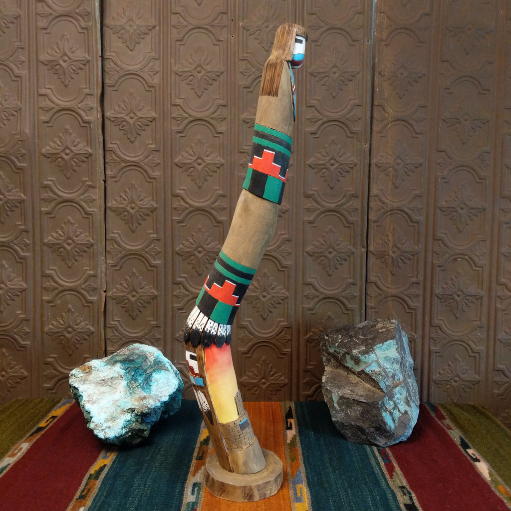 Shalako kachina with vibrant colors, painted feathers, and a pueblo near the base of the piece.