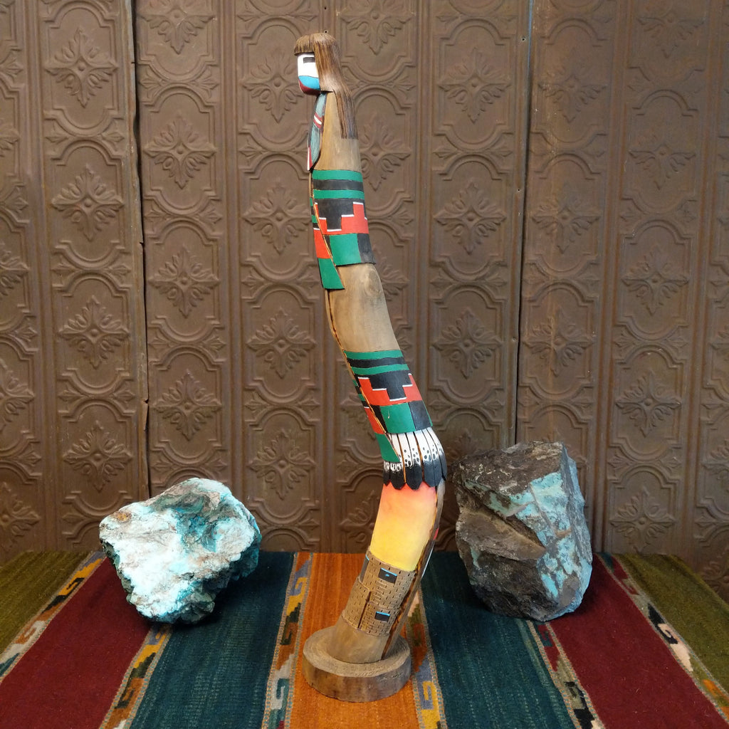 Shalako kachina with vibrant colors, painted feathers, and a pueblo near the base of the piece.