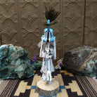 Navajo-made Kokopelli kachina doll by M. C. with blue headdress, peacock feathers, flute, and gray garments.