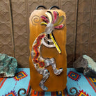Crooks Design metal Kokopelli featuring a brushed silver-toned figure playing a flute, colorful red, yellow, pink, and green accents, and a warm golden-brown metal background.