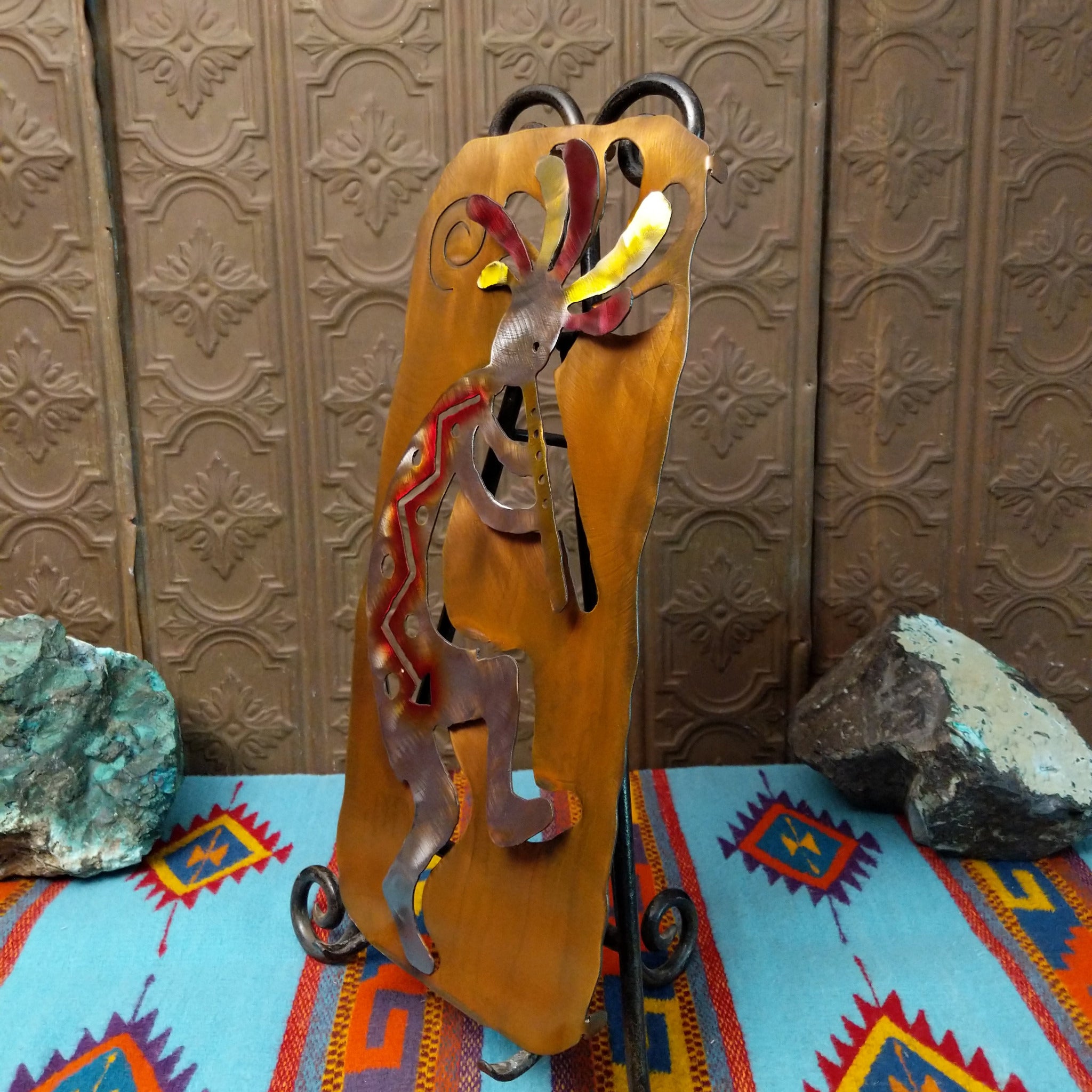 Crooks Design metal Kokopelli featuring a brushed silver-toned figure playing a flute, colorful red, yellow, pink, and green accents, and a warm golden-brown metal background; side view.