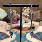 Handcrafted sterling silver arrow dangle earrings by Raymond Coriz featuring round lapis accents, textured arrowheads, and polished silver hooks.