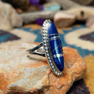 Handcrafted sterling silver ring featuring an elongated marquise-shaped lapis lazuli inlay with diagonal sterling silver geometric accents, framed by a beaded silver border and set on a split sterling silver band; side view.