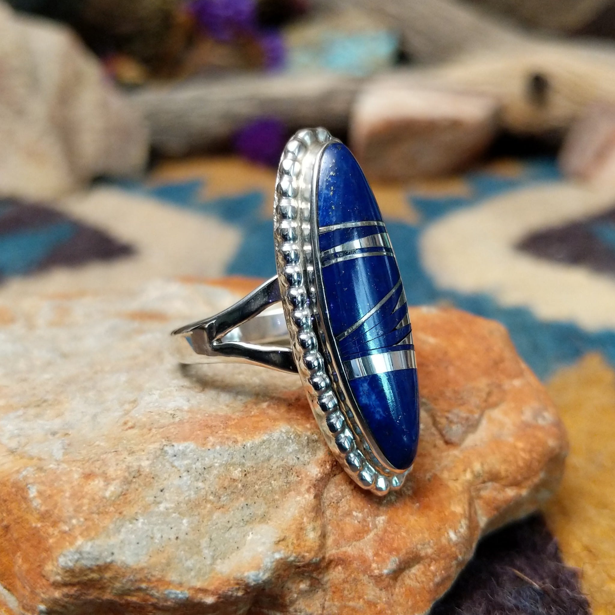 Handcrafted sterling silver ring featuring an elongated marquise-shaped lapis lazuli inlay with diagonal sterling silver geometric accents, framed by a beaded silver border and set on a split sterling silver band; side view.