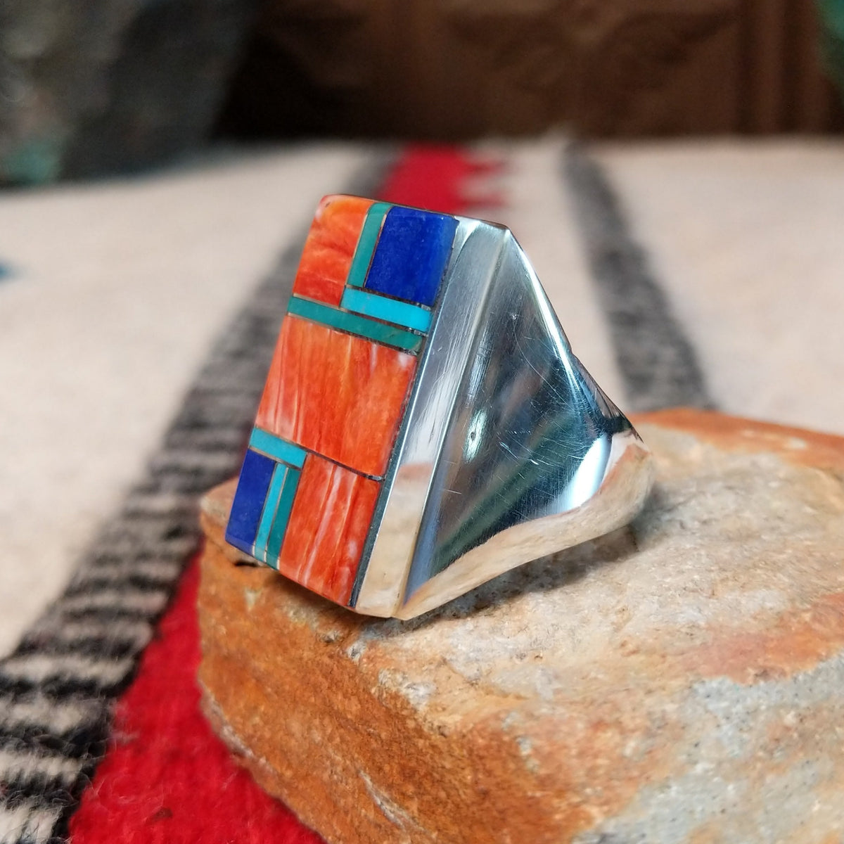 Large multistone inlay ring by Wilbert Grey featuring a rectangular geometric design of orange-red shell, turquoise, and deep blue stones with silver channel accents in a wide polished silver band; side view.